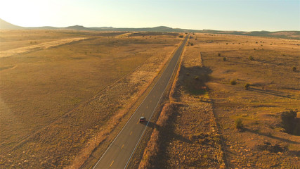 AERIAL: Flying above SUV car driving along empty countryside road at sunset
