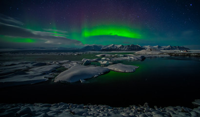 Fototapeta na wymiar A beautiful green aurora borealis dancing over the Jokulsarlon lagoon, Iceland. In the forground is reflection in the lake surface. In the background are the northern lights above the mountains.