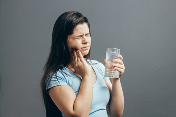 Young woman with sensitive teeth and glass of cold water on gray background
