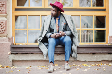 Stylish African American man model in gray coat, jacket tie and red hat sitting against large windows and holding phone at hands.