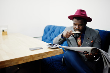 Stylish African American man model in gray jacket tie and red hat drink coffee at cafe and read newspapers.