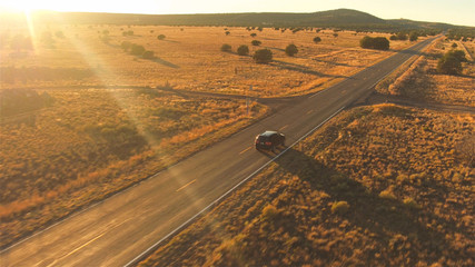 AERIAL: Black SUV car driving along empty country road at golden summer sunset