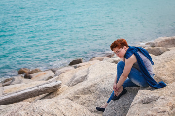 Adult caucasian women sit on rocky beach with blue neckchief relaxing and thinking about something