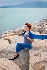 Fototapeta na wymiar Adult caucasian women sit on rocky beach with blue neckchief relaxing and thinking about something
