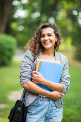 Pretty cheerful latin american student smiling at camera carrying notebook on campus at college - obrazy, fototapety, plakaty