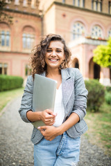 Portrait of young beautiful woman student walking in the park using mobile phone holding laptop...
