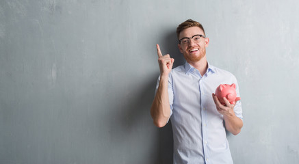 Young redhead man over grey grunge wall holding piggy bank surprised with an idea or question...