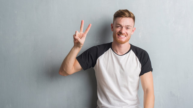 Young redhead man over grey grunge wall showing and pointing up with fingers number two while smiling confident and happy.