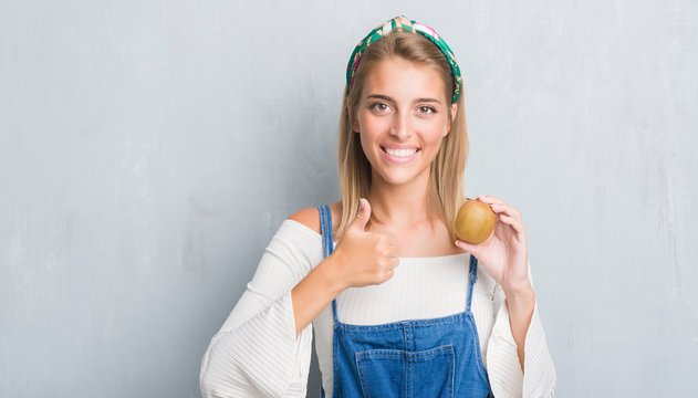 Beautiful young woman over grunge grey wall holding fresh kiwi happy with big smile doing ok sign, thumb up with fingers, excellent sign