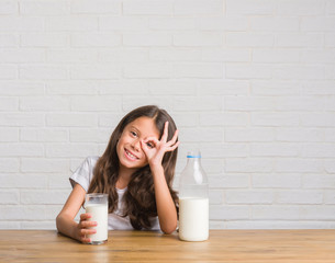 Obraz na płótnie Canvas Young hispanic kid sitting on the table drinking a glass of milk with happy face smiling doing ok sign with hand on eye looking through fingers
