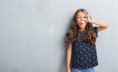 Naklejka premium Young hispanic kid over grunge grey wall doing ok gesture shocked with surprised face, eye looking through fingers. Unbelieving expression.