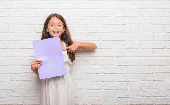 Young hispanic kid over white brick wall holding pink paper sheet with surprise face pointing finger to himself