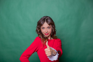 Smiling attractive woman in red dress is posing with hand on hip and showing thumb up on green turquoise background.pretty happy young businesswoman is satisfied, joyful.Copy space - Powered by Adobe