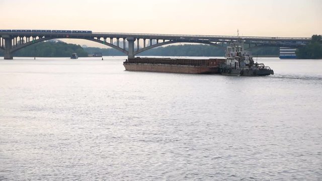 A small ship tows a barge with a cargo on the river in the direction of the bridge. Transportation by water.