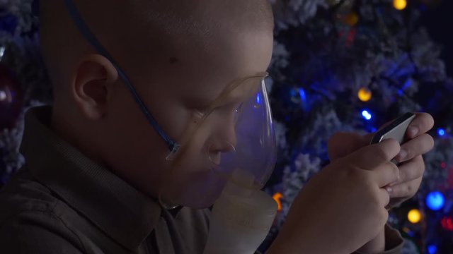Unhappy boy with shaved head inhales vapors containing medicines for inhaler to stop cough. Child is looking information about his illness in smartphone.  Respiratory medicine. Bronchitis problem