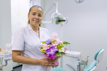Female dentist smiling, with a bouquet of flowers, in dental office. National dentist's day.