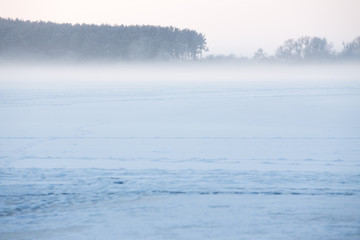 Christmas winter landscape of the frozen lake covered with snow on a cold winter day and the forest in thr mist 
