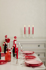 Merry Christmas and Happy New Year! Table setting - red and pink dishes, holiday knitted decor - Santa Claus knitted hats on the bottle with wine, red candles.