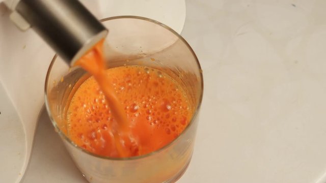 Carrot juice pours from a juicer into a clear glass tumbler on the white background