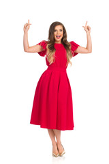 Happy Fashion Woman In Red Dress And Gold High Heels Is Looking At Camera, Talking And Pointing Up