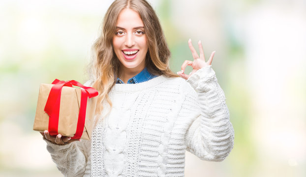 Beautiful young blonde woman holding gift over isolated background doing ok sign with fingers, excellent symbol