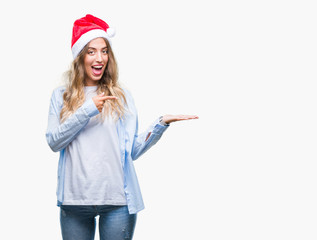 Beautiful young blonde woman wearing christmas hat over isolated background amazed and smiling to the camera while presenting with hand and pointing with finger.