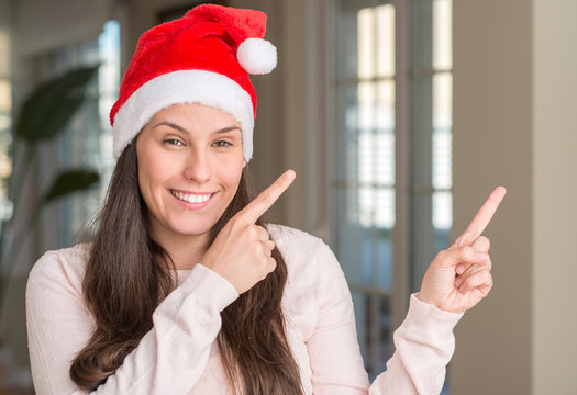 Beautiful young woman wearing Santa Claus hat at home smiling and looking at the camera pointing with two hands and fingers to the side.