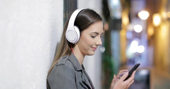 Side view portrait of a happy girl relaxing listening to music from smart phone in the night in the street