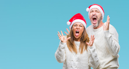 Obraz premium Middle age hispanic couple wearing christmas hat over isolated background crazy and mad shouting and yelling with aggressive expression and arms raised. Frustration concept.