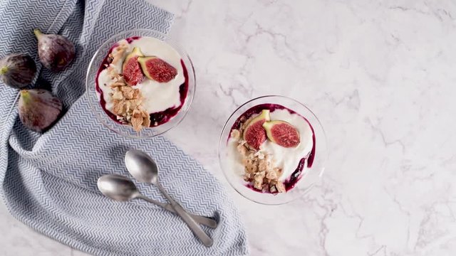 Figs pudding parfait with yogurt, blueberry jam, figs, hazelnut, and cookies in glass on white marble background. Flatlay top view.