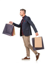 side view of adult male shoppper in jacket walking with colorful paper bags isolated on white