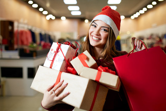 Happy girl shopping gifts in mall on christmas sale. New year holidays shopping idea concept. Smiling woman with colorful paper presents bags and gift boxes wearing christmas hat in store or shop.