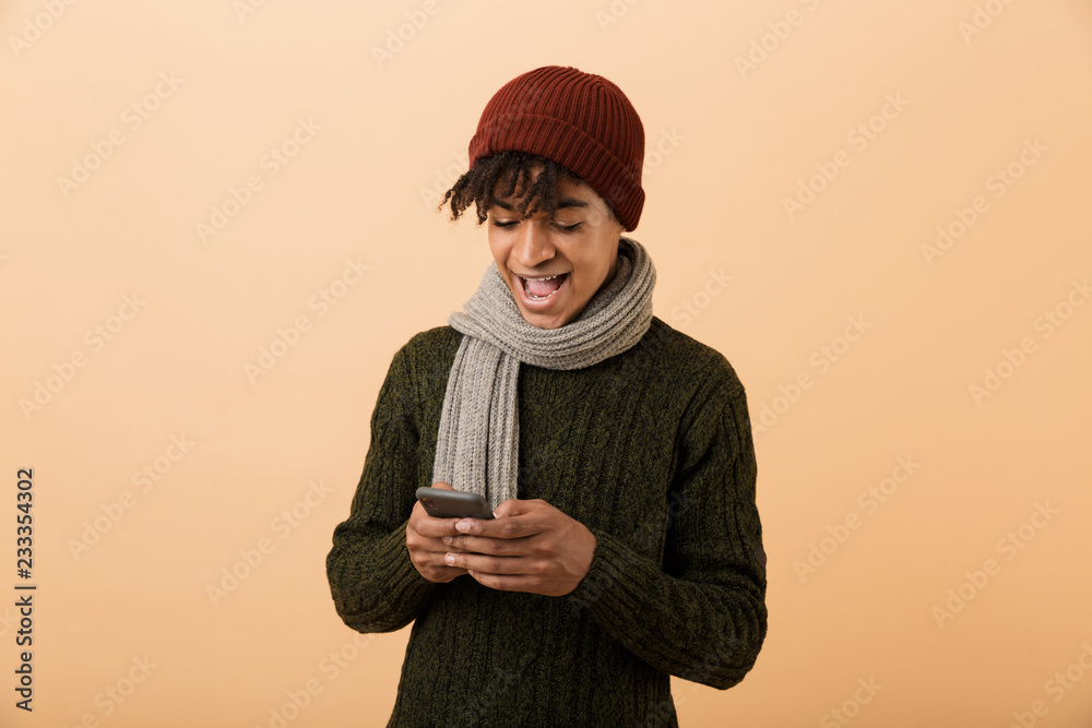 Poster portrait of positive teen man wearing hat and scarf using cell phone, isolated over yellow backgroun