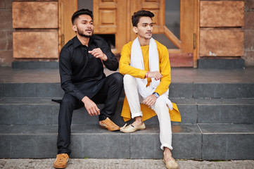 Two indian stylish mans friends in traditional clothes posed outdoor and sitting on stairs.