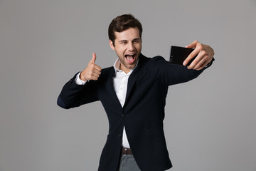 Image of joyous businessman 30s in formal suit showing thumb up while taking selfie photo on cell phone, isolated over gray background