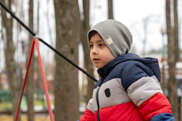 Obraz premium A boy in a jacket and hat is playing on the playground in the park. He deftly climbs the rope attraction.