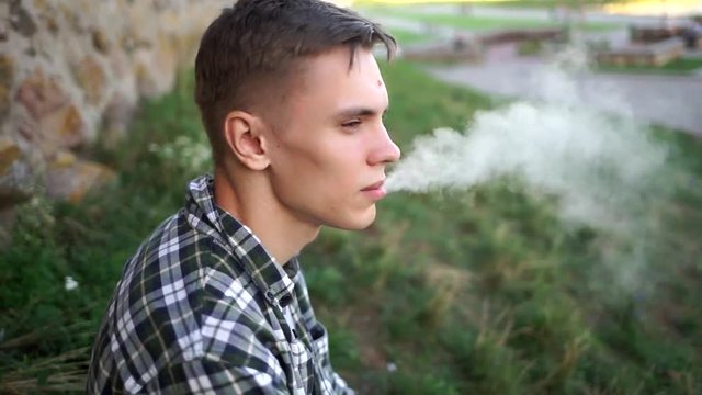 Beautiful model smokes electronic cigarette. Cute guy smiles and blows smoke from vaping. A man in a plaid shirt near the wall smokes an electronic cigarette. Handsome guy blowing smoke in the cell.