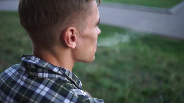 Beautiful model smokes electronic cigarette. Cute guy smiles and blows smoke from vaping. A man in a plaid shirt near the wall smokes an electronic cigarette. Handsome guy blowing smoke in the cell.