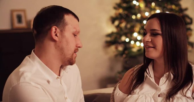 Husband and his pregnant wife drinking delicious tea in mugs while sitting on cozy comfy sofa in living room decorated for Christmas. Woman is waiting for a baby, man is touching her belly with love
