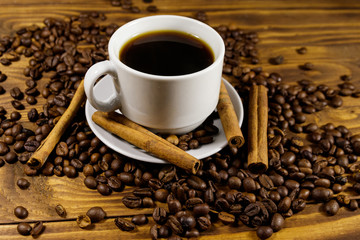 Cup of coffee, roasted coffee beans and cinnamon sticks on wooden table