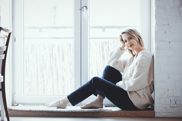 young girl dressed in a sweater and jeans is sitting at the threshold of the window at home
