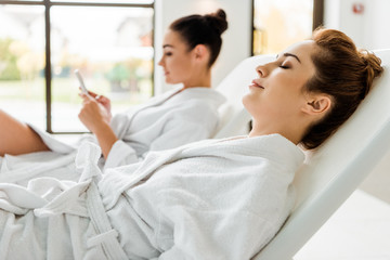 side view of young women in bathrobes relaxing on sunbeds in spa