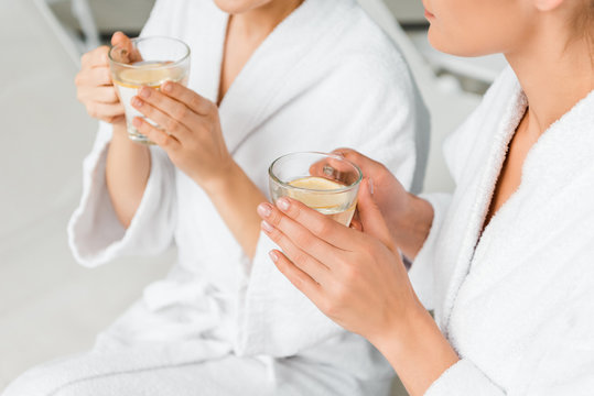 cropped shot of young women in bathrobes holding cups with herbal tea and lemon while resting in spa