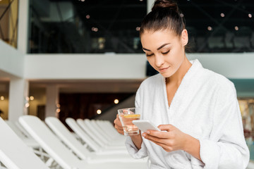 young woman in bathrobe holding cup of herbal tea and using smartphone in spa