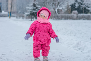 Fototapeta na wymiar Baby in winter overalls wrapped in a scarf walks on a snow-covered street and sculpts from snow.