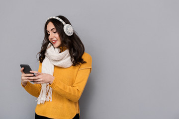 Portrait of happy woman wearing ear muffs and scarf holding mobile phone, isolated over gray background