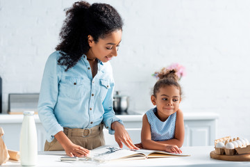african american mother and daughter reading recipe book in kitchen