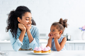 cheerful african american mother and daughter resting chins on hands and looking at each other, cupcakes on table in kitchen