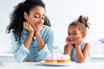 happy african american mother and daughter resting chins on hands near cupcakes on table in kitchen