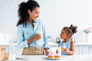 african american mother and daughter standing with glasses of milk and cupcakes in kitchen, looking at each other